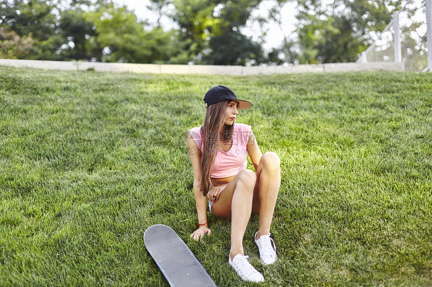 旁边放着滑板的年轻女人坐在草地上图片
