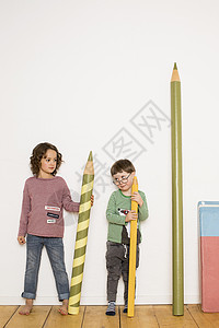 年轻女孩和男站立拿着巨型铅笔文具贴在他旁边的墙上图片