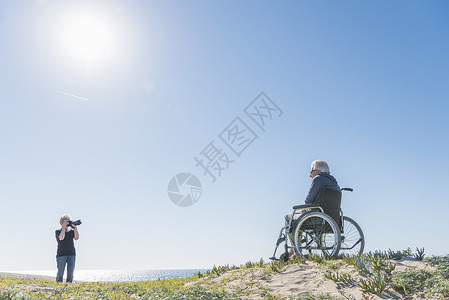 老年女人给沙丘上给坐轮椅的丈夫拍照图片