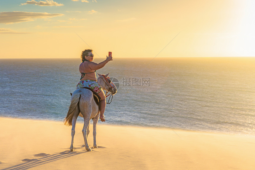 妇女骑马在海滩上用智能手机拍照图片