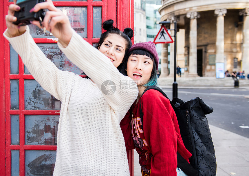 两位年轻时尚女人在红色电话亭旁边用手机自拍英国伦敦图片