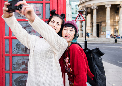 两位年轻时尚女人在红色电话亭旁边用手机自拍英国伦敦图片