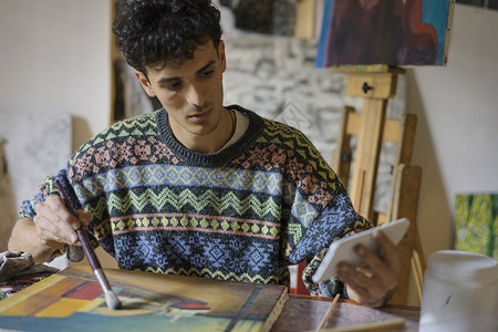 男艺术家在艺术家工作室画画布时看着智能手机图片