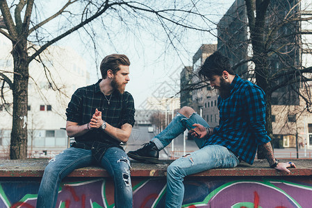 坐在围墙上看着智能手机的两位年轻时尚潮男图片