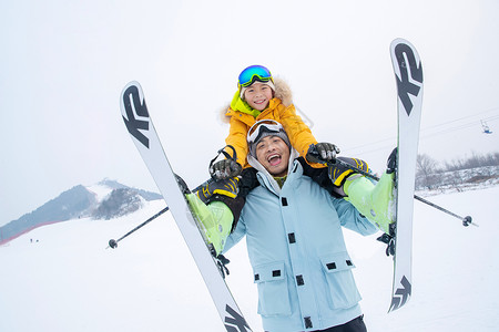 滑雪场上拿滑雪杖的快乐男孩骑在父亲脖子上高清图片