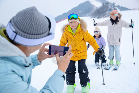 逃避现实周末活动厚衣服滑雪场上父亲用手机给孩子们和妻子拍照图片