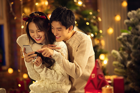 漂亮圣诞树圣诞节东方人男朋友幸福的年轻伴侣在家玩手机背景