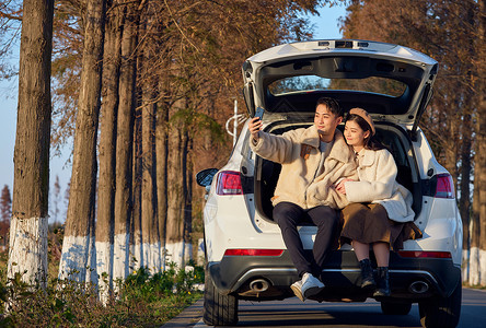 年轻情侣自驾出游坐在汽车后备箱拍照背景图片