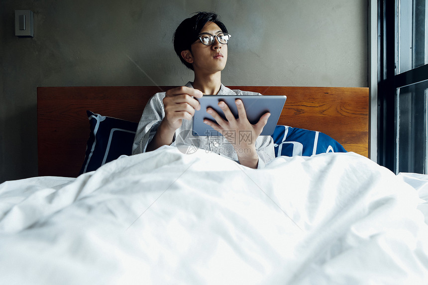 穿睡衣躺在床上食用平板电脑的年轻男性图片