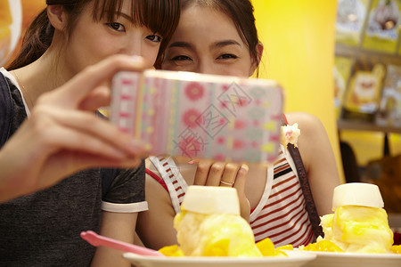 两个女游客坐在放着刨冰的桌边用手机自拍图片