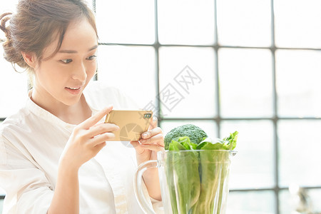 年轻女子拿着手机拍搅拌机里的蔬菜图片