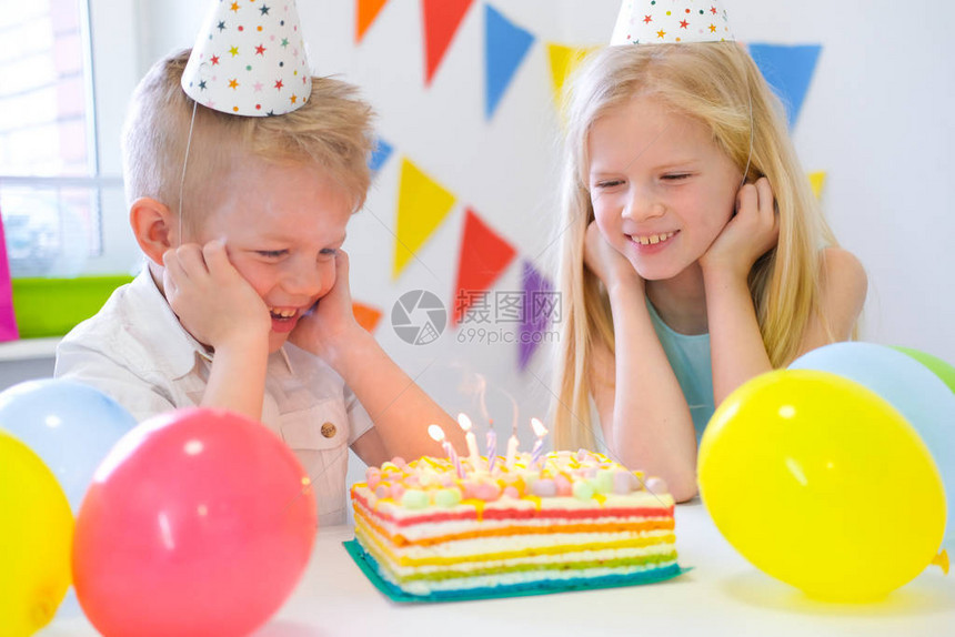 两个金发的caucasian男孩女看着生日彩虹蛋糕点着蜡烛和在生日派对上许愿图片