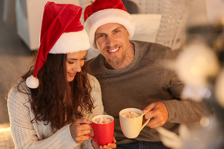 穿着圣诞帽子的可爱小情侣在家庆祝圣诞节时喝热巧克图片