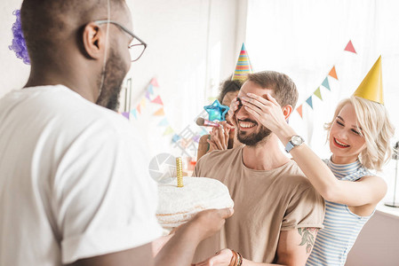 快乐的朋友遮着年轻男人的眼睛用生日蛋糕向他问好图片