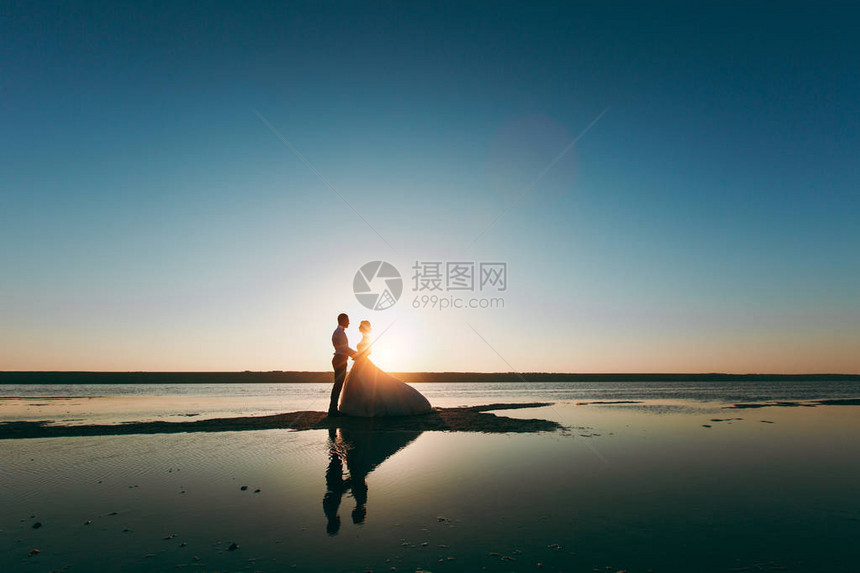 年轻的新娘和新郎身着长裙在海边的海岸线上的剪影图片