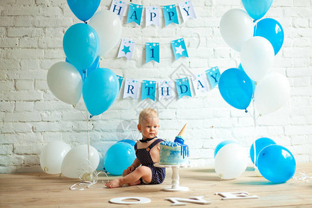 一岁的男孩在很多蓝色和白色的气球节日蛋糕和砖墙和地板上的铭文中庆祝他图片