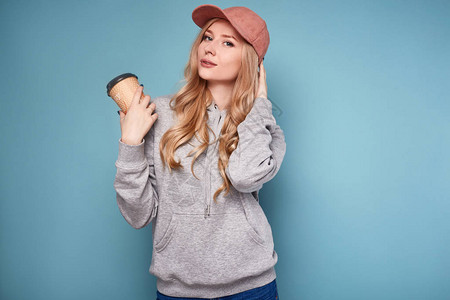 一个穿着粉红棒球帽喝咖啡和运动衫的漂亮金发美女肖像在工作室图片