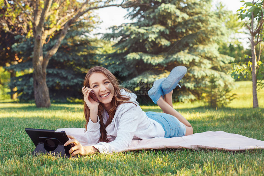 年轻美丽的白种人少女在公园外用手机交谈的画像带着数字平板电脑和手机的微笑女人躺在公园的草图片