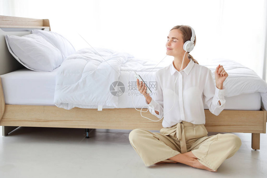 年轻漂亮的白种女人穿着带耳机的白色休闲服她在卧室里用手机或音乐播放器听音乐或收音机图片