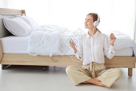 年轻漂亮的白种女人穿着带耳机的白色休闲服她在卧室里用手机或音乐播放器听音乐或收音机背景图片