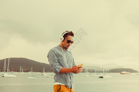 留着胡须的男人穿着太阳镜衬衫和短裤在海边听音乐发短信图片