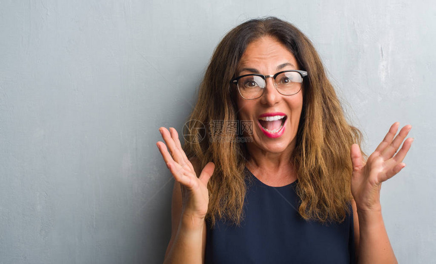 戴眼镜的中年西班牙裔女在灰色墙上非常高兴和奋图片