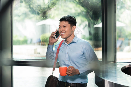 一位英俊开朗的亚洲年轻人在商务楼端着咖啡打着电话图片