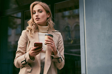 穿着海沟大衣的美丽散心金发美女喝着咖啡到外面去手机自信地在户图片