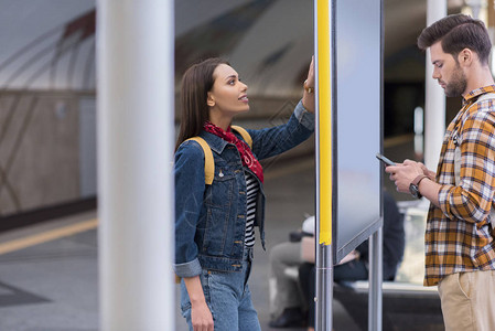有智能手机的男游客及其女友在地铁站看信息板时观看其侧面图片