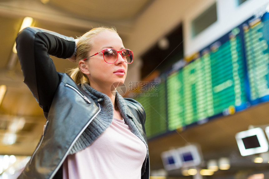 穿着随意的年轻时尚女旅客在机场候机楼大厅的登机柜台前检查出发板航班时刻表显示在背景中模糊图片