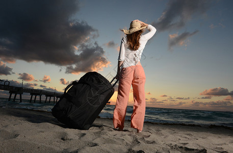 女人拖着行李箱带着沙滩帽在沙滩看日落图片