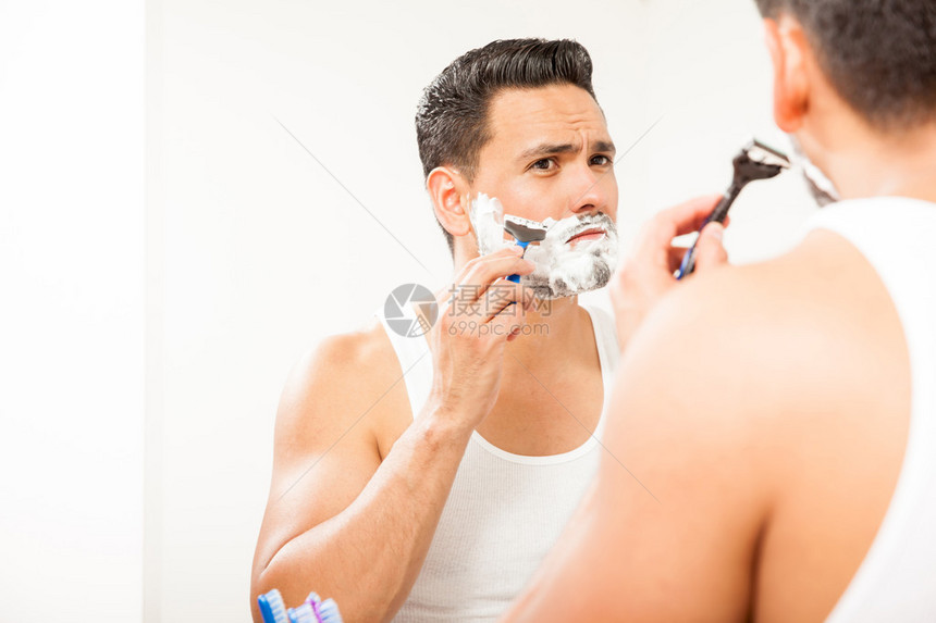 一个有魅力的西班牙青年男子的肖像利用剃刀在浴室镜子前刮图片