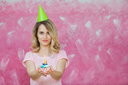 穿着生日帽子的漂亮金发美女拿着蛋糕和一根蜡烛在粉红背景上图片