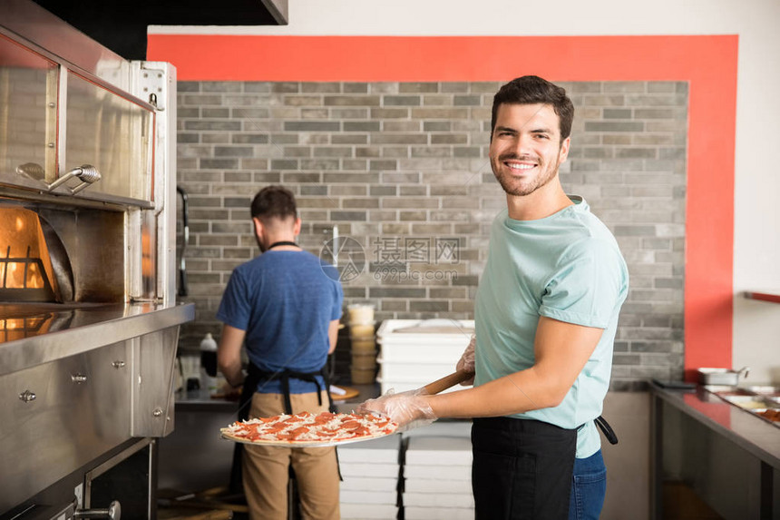 穿着黑色围裙的微笑厨师把披萨放进烤炉里在餐厅图片