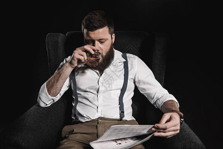 时髦的男人喝威士忌和看报纸而坐在扶椅上被隔离在黑图片