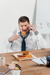 超重量超重商人在工作场所吃垃圾食品时使用图片