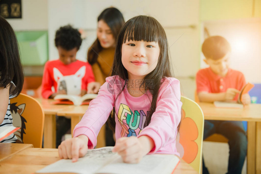 读一本书的亚洲女孩对着镜头微笑一排多民族小学生在学校的教室里读书复古图片