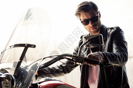 穿着太阳眼镜和皮夹克的帅气英俊年轻男子坐在摩托车上背景图片
