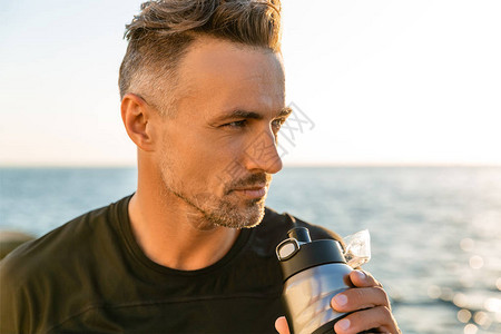 英俊成年男子在日出前从海边健身水瓶中取来饮用图片