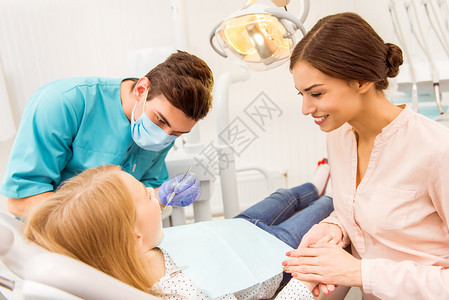 在牙科诊所与母亲一起检查年轻病人的儿科牙医图片