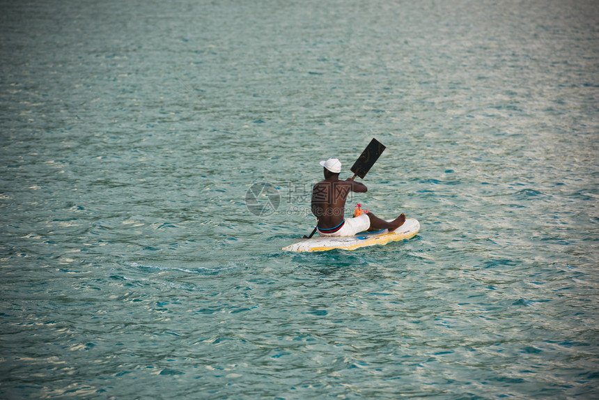 年轻的塞舌尔男子在海中冲浪板上卖水果图片