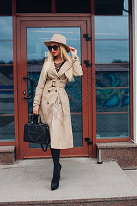 穿着时髦的米色风衣米色帽子和黑色太阳镜的华丽自信金发模特的库存照片图片
