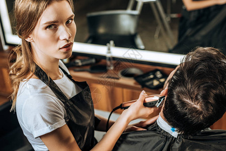 利用电子剪发机和在美容院与客户合作时观看照相机的年轻理发师的高图片