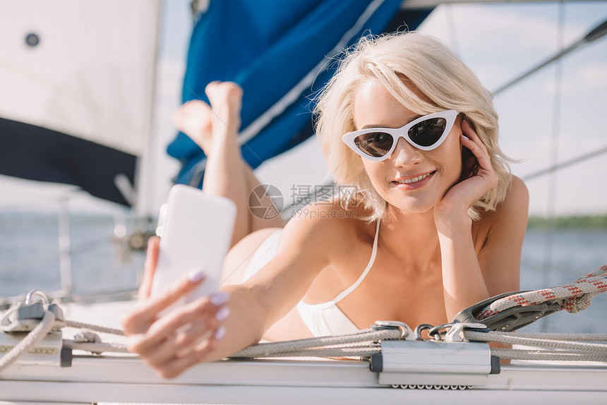 穿着比基尼和太阳镜的有笑容的迷人女躺在游艇上时用图片