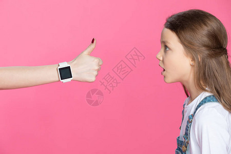 惊吓的小女孩的侧面景观看着有智慧的手表的女人拇指在粉图片