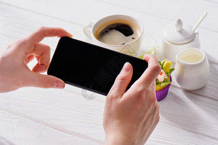 手持智能手机空白屏幕咖啡杯奶油和甜松饼在木头表面图片