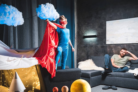 英雄出少年儿子穿着红色超级英雄服装在家中玩父亲背景
