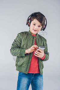 有耳机和智能手机的可爱男孩看着在灰色上图片
