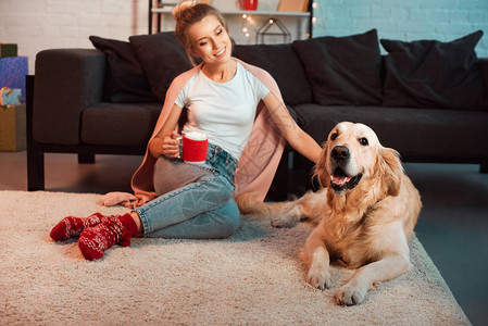 快乐的金发年轻女子狗坐在毛地毯上喝着热可圣诞节图片
