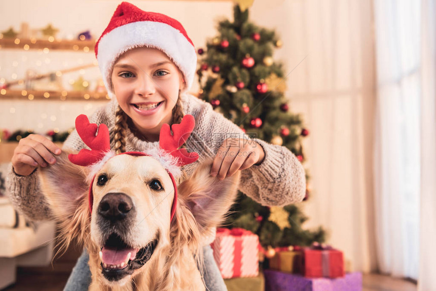 穿着圣塔帽子的笑着孩子带着鹿角的猎犬在圣诞树附近图片